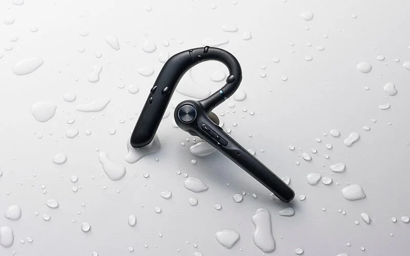 Black ear-hook Bluetooth headset with water droplets on a white background, indicating waterproofness