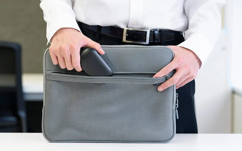 Person storing a black gadget in the mesh pocket of a grey laptop sleeve, demonstrating portability