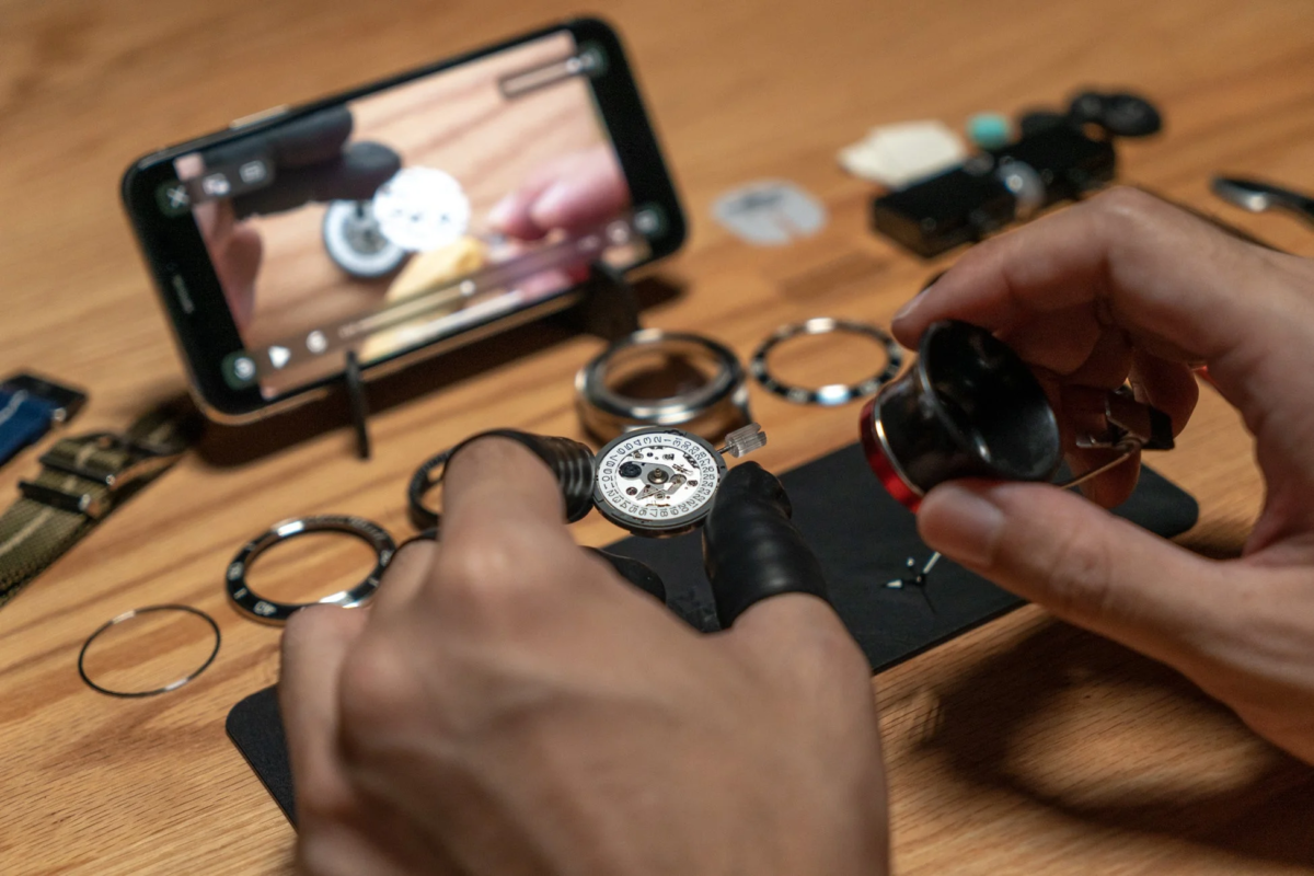 Hand assembling a watch movement while watching a tutorial on a smartphone