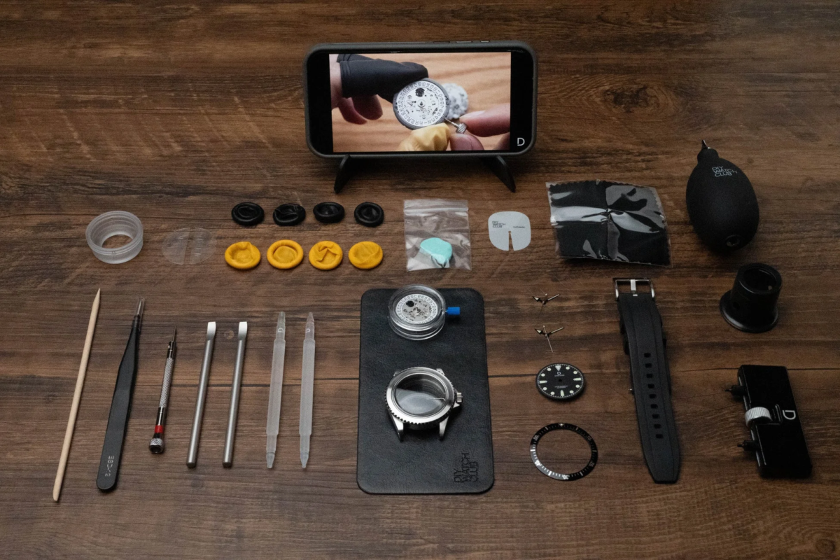 DIY watch kit tools and components laid out on a wooden table, with a smartphone displaying a tutorial video