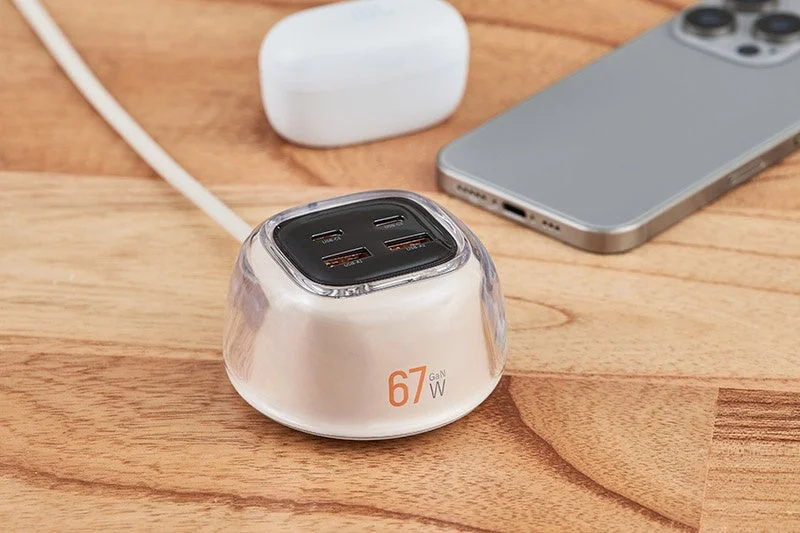 A 67W GaN multi-port USB charger on a wooden surface, with a smartphone and wireless earbuds nearby.