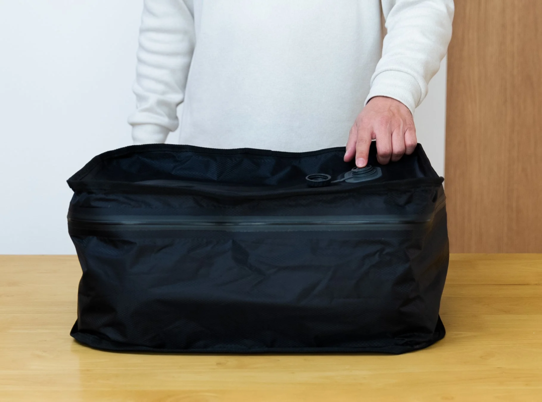 A person's hand pressing a valve on a black compression bag on a wooden table, suggesting ease of use for packing.