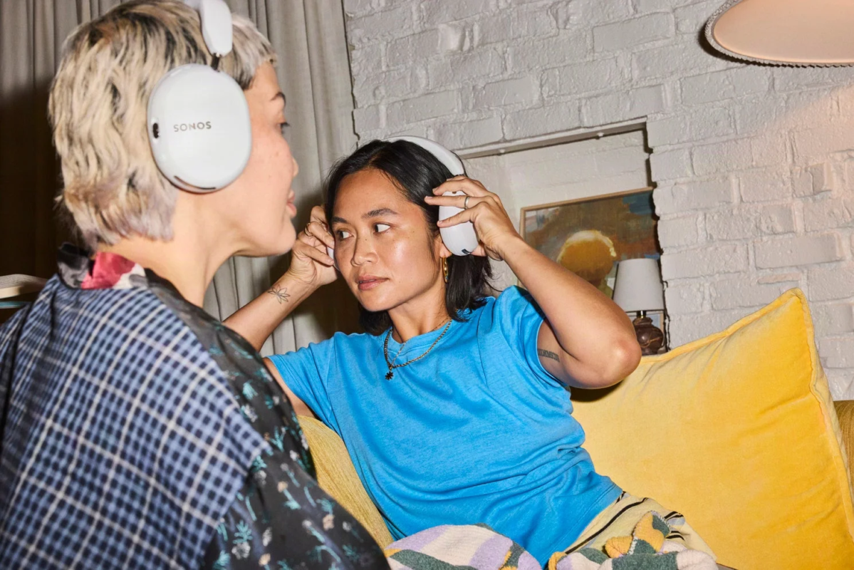 Two women wearing headphones, one with white Sonos headphones, relaxing on a yellow sofa